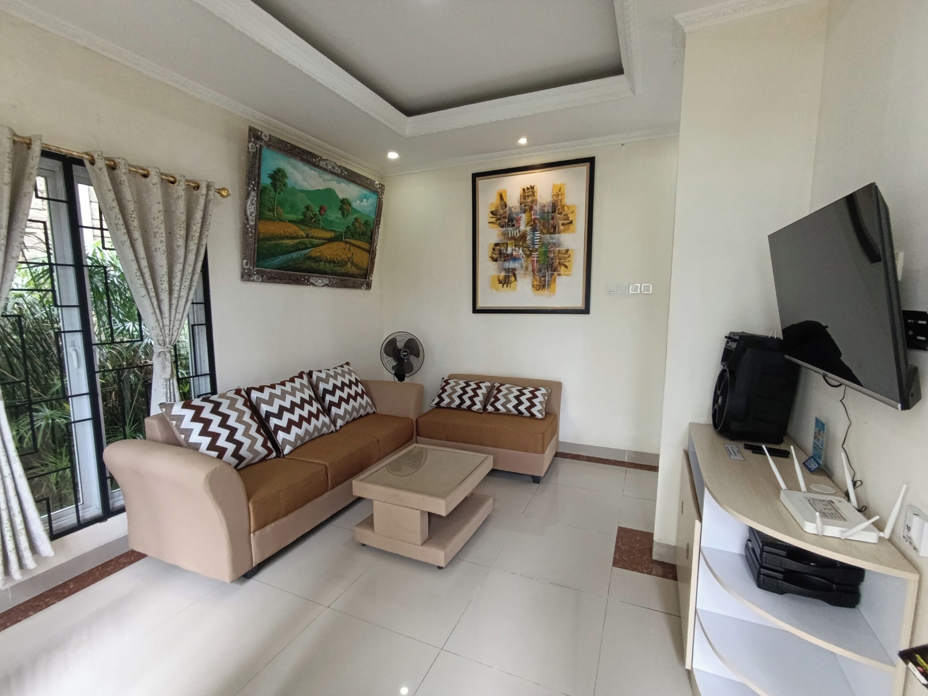 A living room with a beige sectional sofa, a coffee table, a TV, and a fan. There is a painting on the wall and a window with curtains.