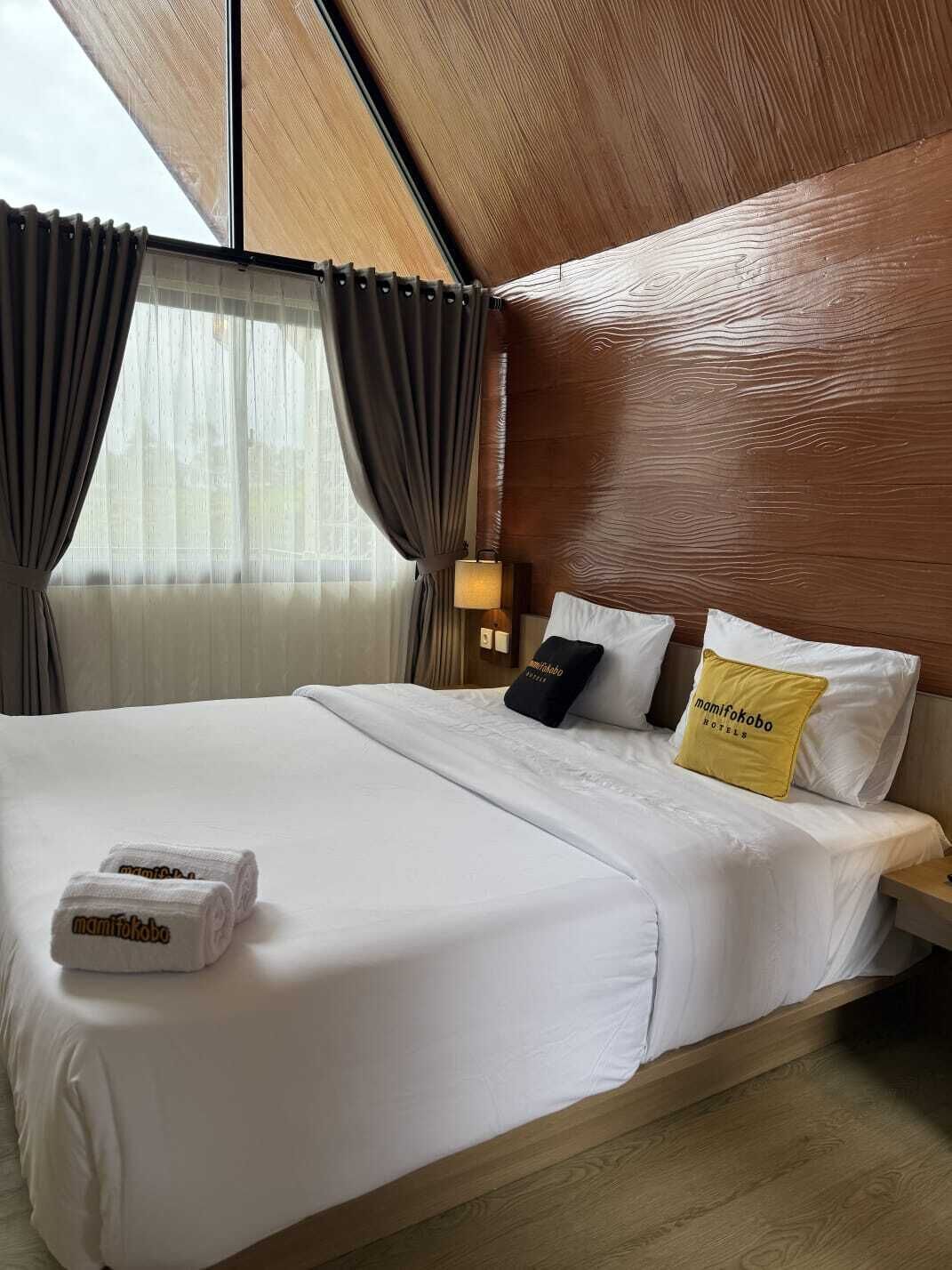 A hotel room with a large bed, two pillows, a nightstand, a lamp, and a window with curtains.