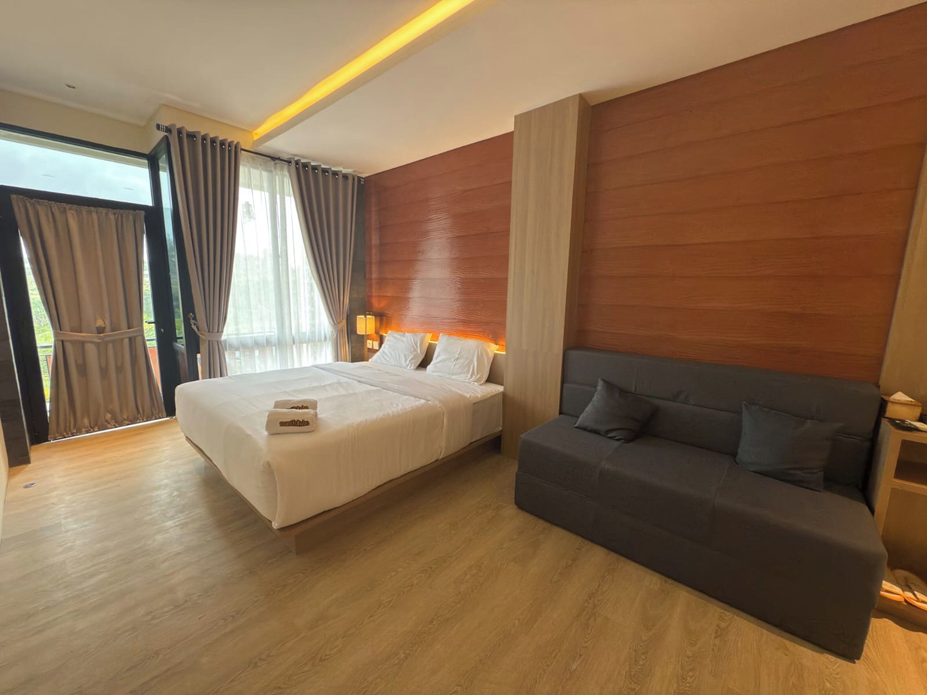 Una habitación de hotel con cama, sofá, suelo de madera y un ventanal con cortinas.