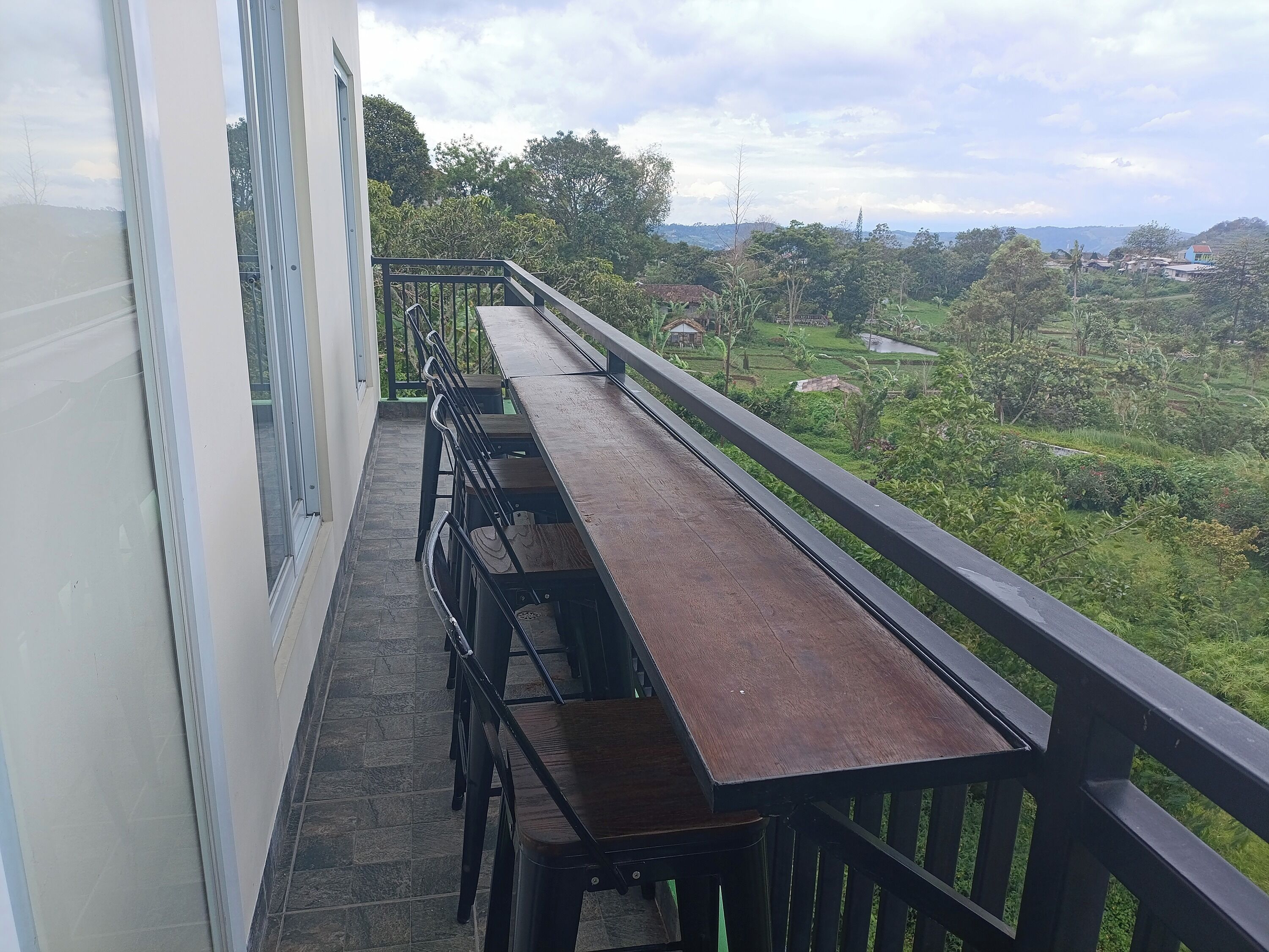 A balcony with a long wooden table and chairs, overlooking a lush green landscape with distant buildings.