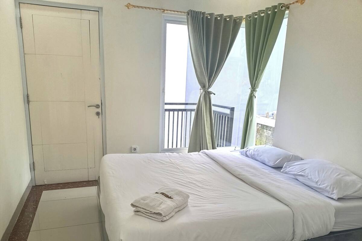 Un llit ben fet amb roba blanca i una tovallola doblegada, una finestra amb cortines verdes i una porta blanca.