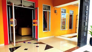 A hotel entrance with a red and yellow facade, a tiled floor with geometric patterns, and a visible room with a bed and a window. - Esemka Hotel and Convention Kotabumi (Kali Balangan)
