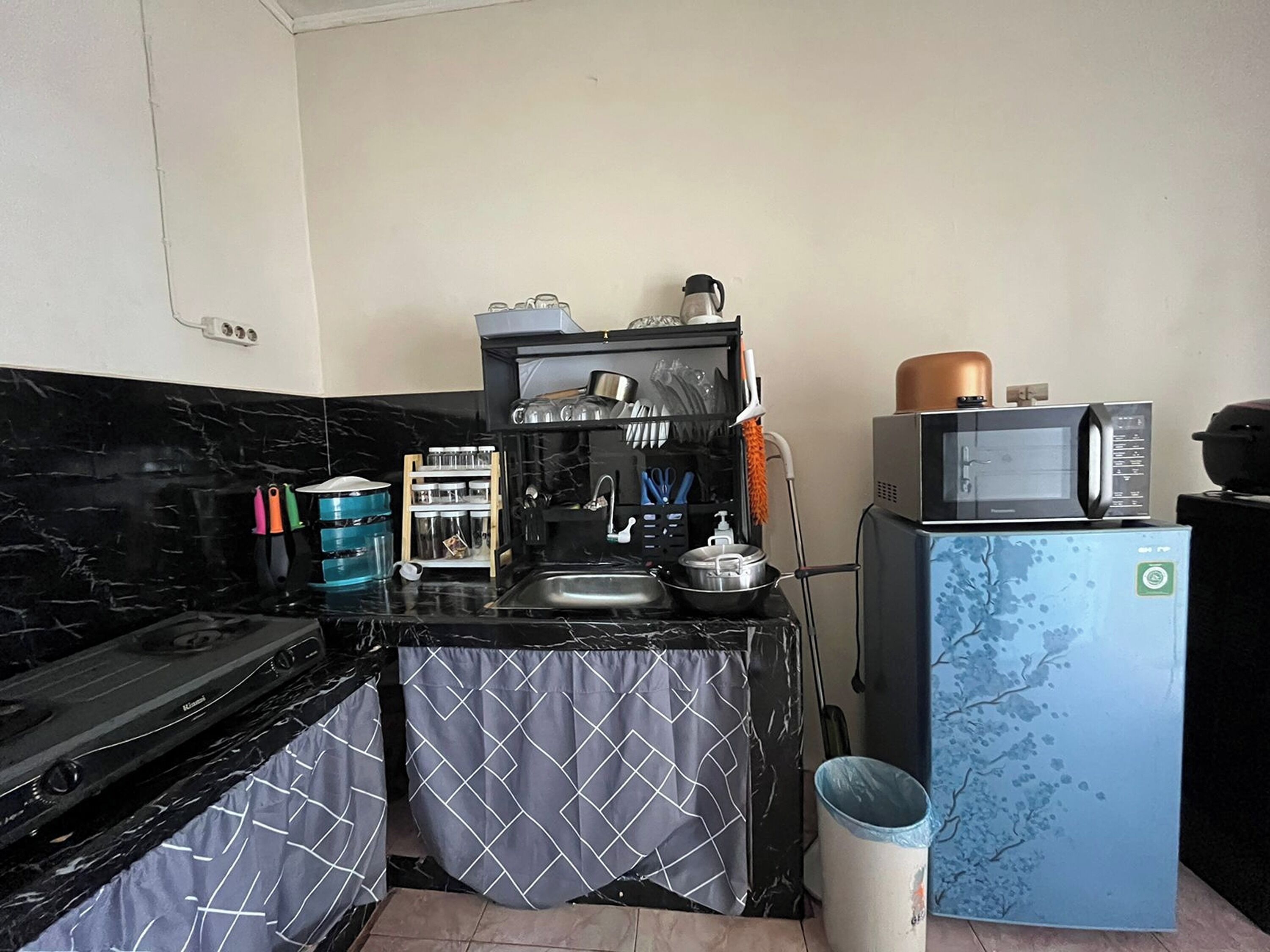 Une cuisine de taille réduite, équipée d’une cuisinière, d’un évier et d’un four à micro-ondes. On trouve des placards au-dessus de l’évier et une poubelle sur le côté.