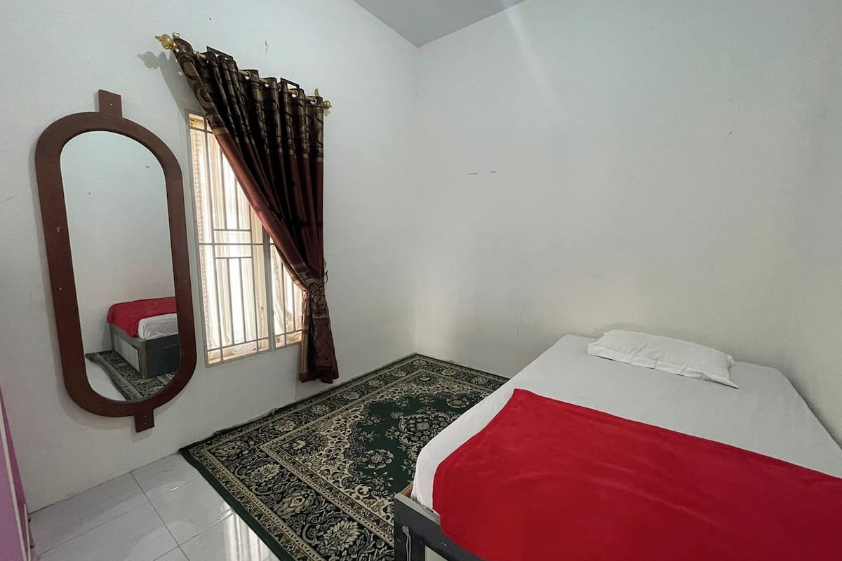 Una habitación pequeña y limpia con cama, espejo, ventana con cortinas y una alfombra estampada.