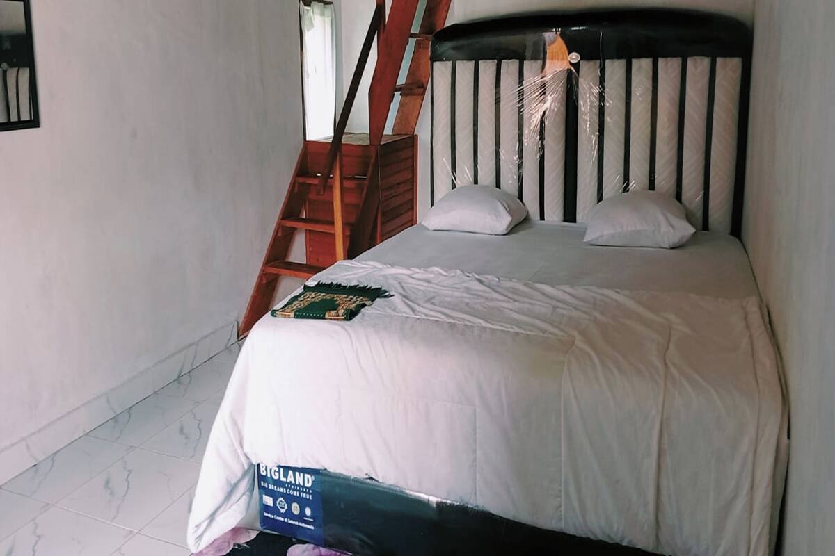 Ein Schlafzimmer mit einem Bett, Kissen, einer Decke und einem Schild auf dem Bett. Im Hintergrund ist eine Holztreppe zu sehen.