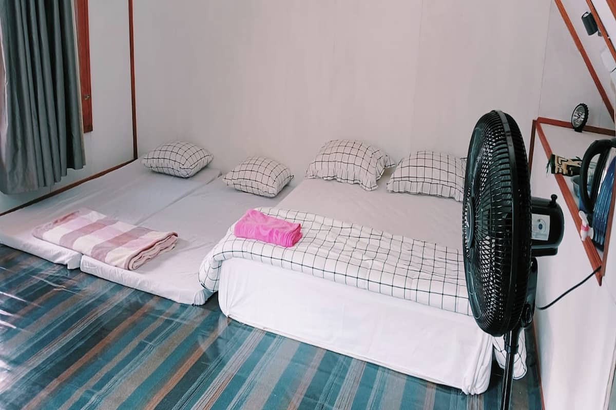 Ein Einzelbett mit karierter Bettdecke, ein pinkfarbenes Handtuch und mehrere Kissen. Ein Standventilator befindet sich rechts vom Bett.
