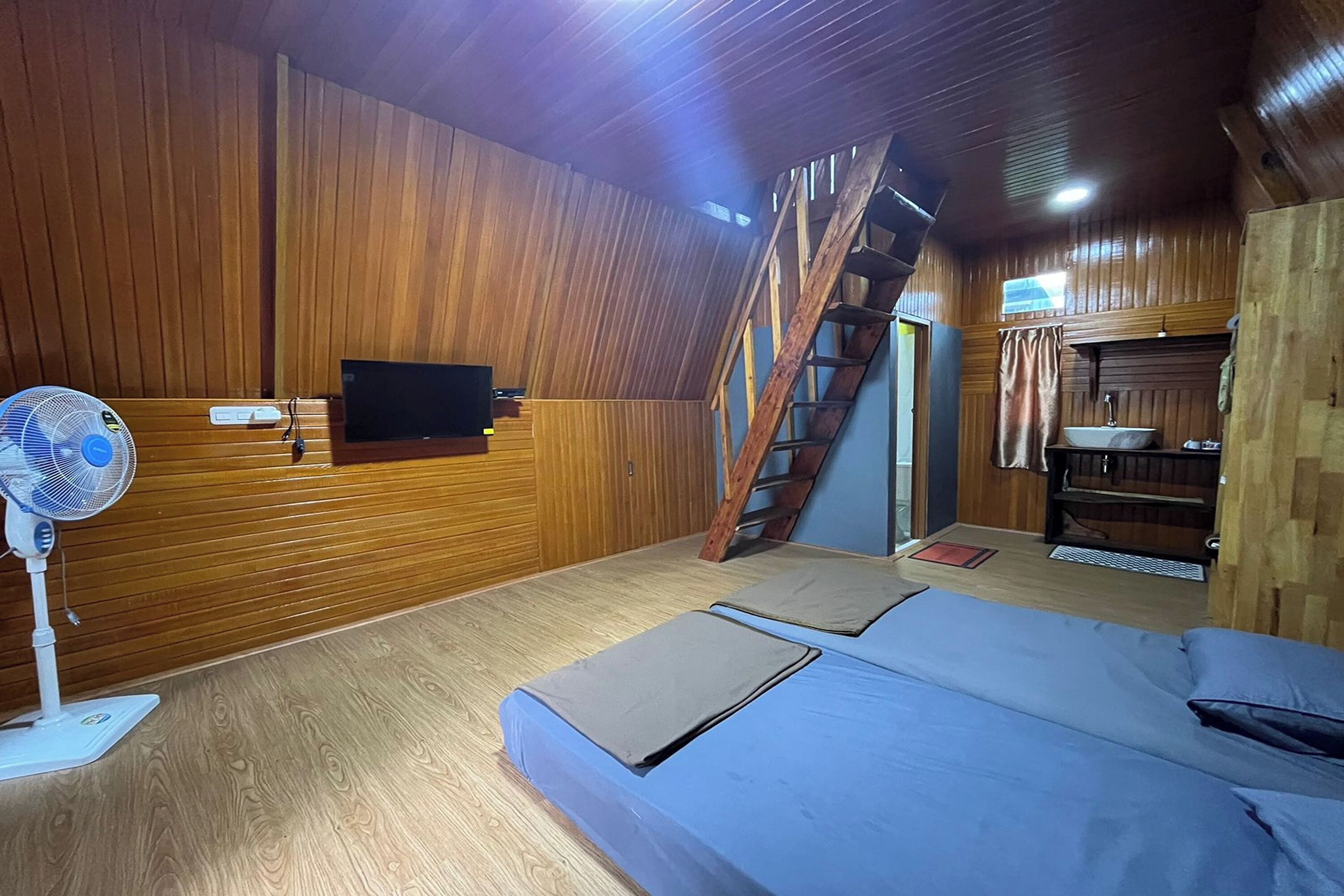 A room with a bed, a fan, a TV, and a wooden staircase.