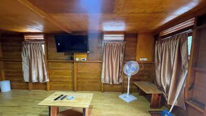 A wooden cabin interior with a wooden table, a bench, a fan, and a television mounted on the wall. - Villa Tapian Banda (Bukittinggi)