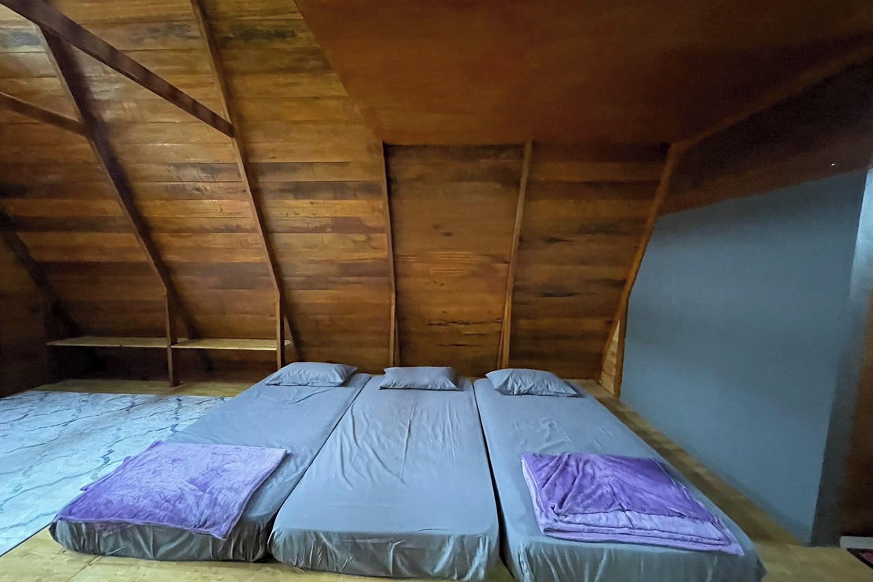 A room with three single beds aligned against a wooden ceiling, a small shelf, and a plain wall.