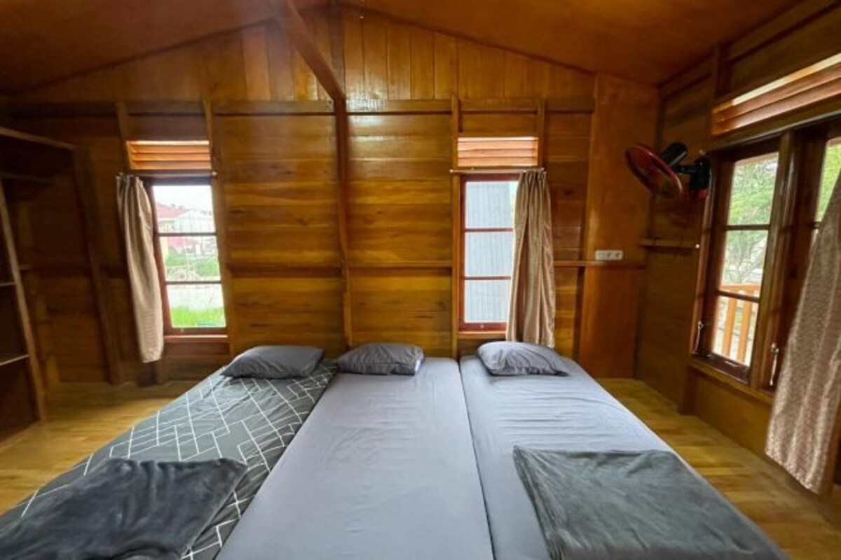 A cabin with a long, narrow room featuring two beds with grey bedding and a central bed with a white sheet. There are wooden walls and a window with curtains.