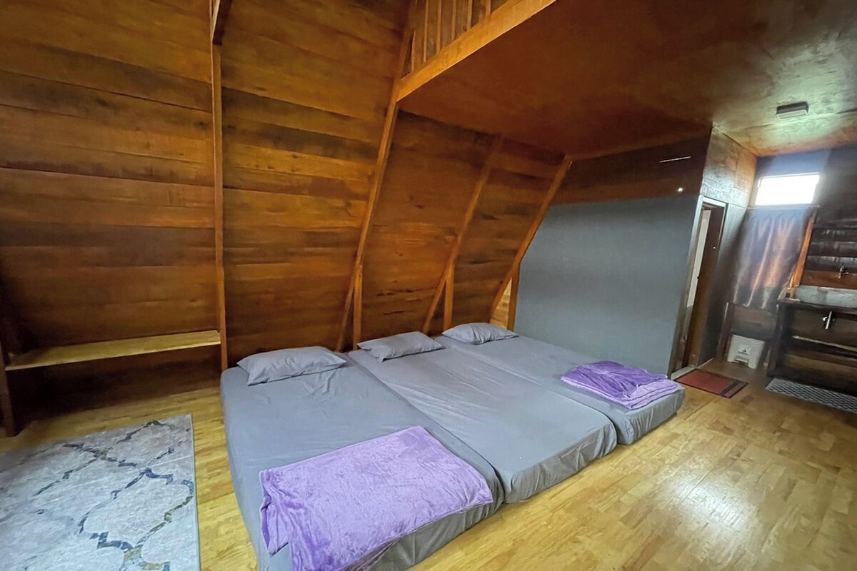 A room with a sloped ceiling, wooden walls, a bed with grey bedding, and a small wooden table.