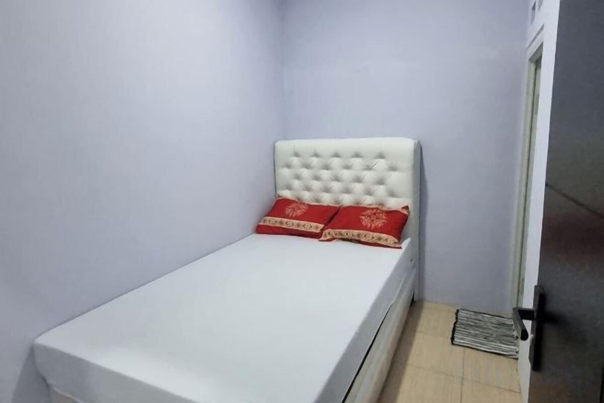 Ein kleines Zimmer mit einem Einzelbett, einem weißen Kopfteil und roten Zierkissen.