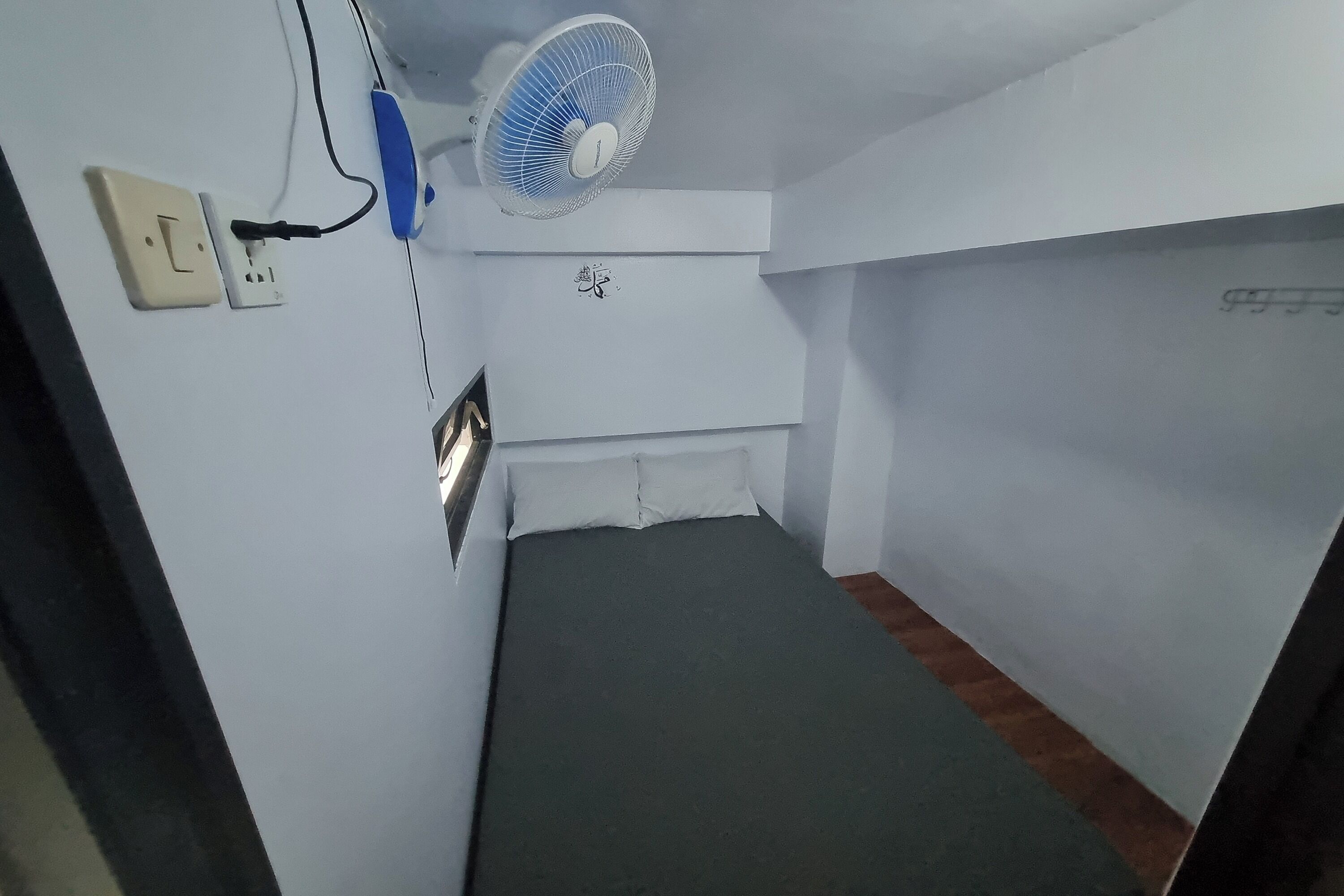 A small room with a bed, a wall-mounted air conditioner, a ceiling fan, and a power outlet.