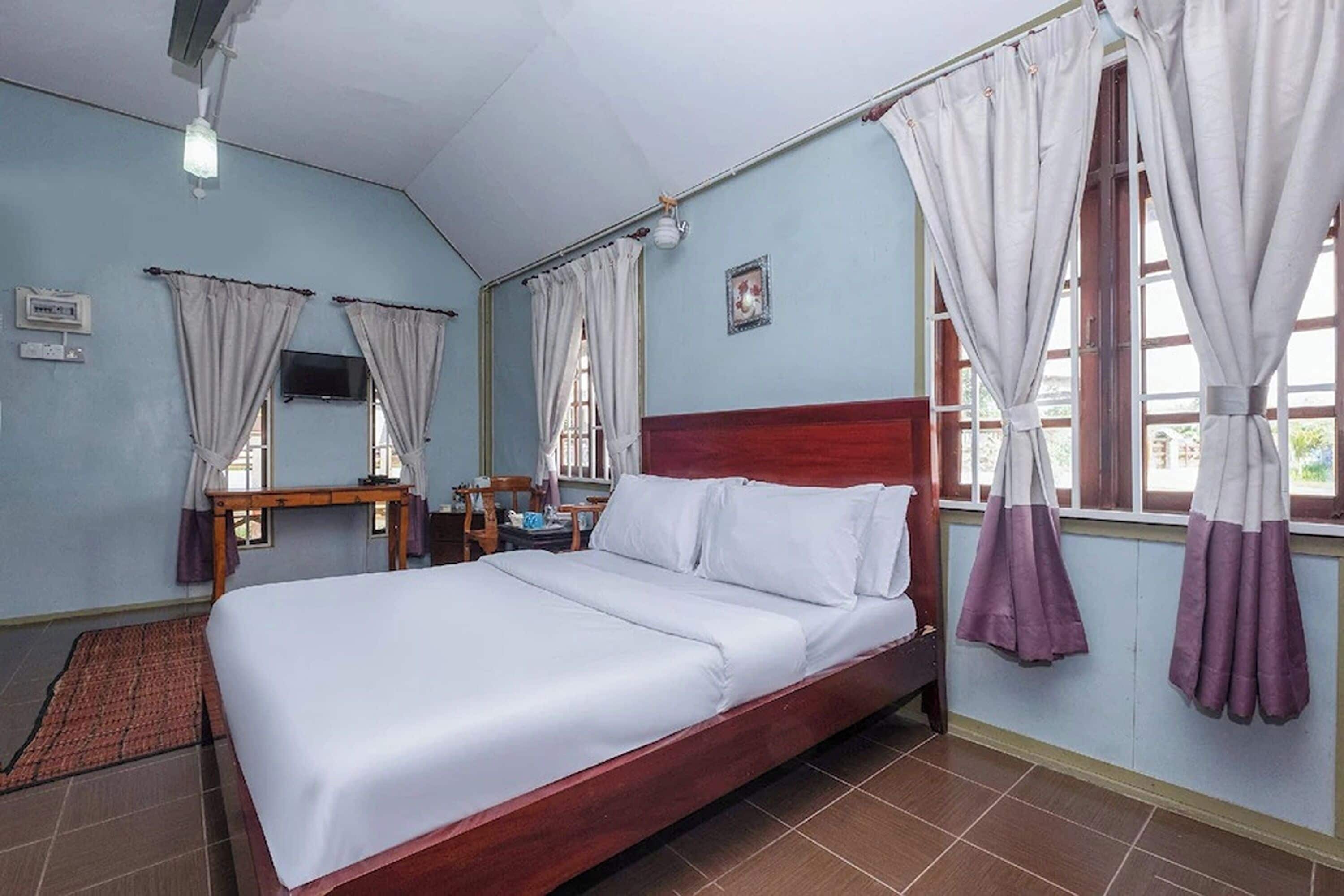A hotel room with a wooden bed, a desk, and a chair. There are curtains on the windows and a television on the wall.
