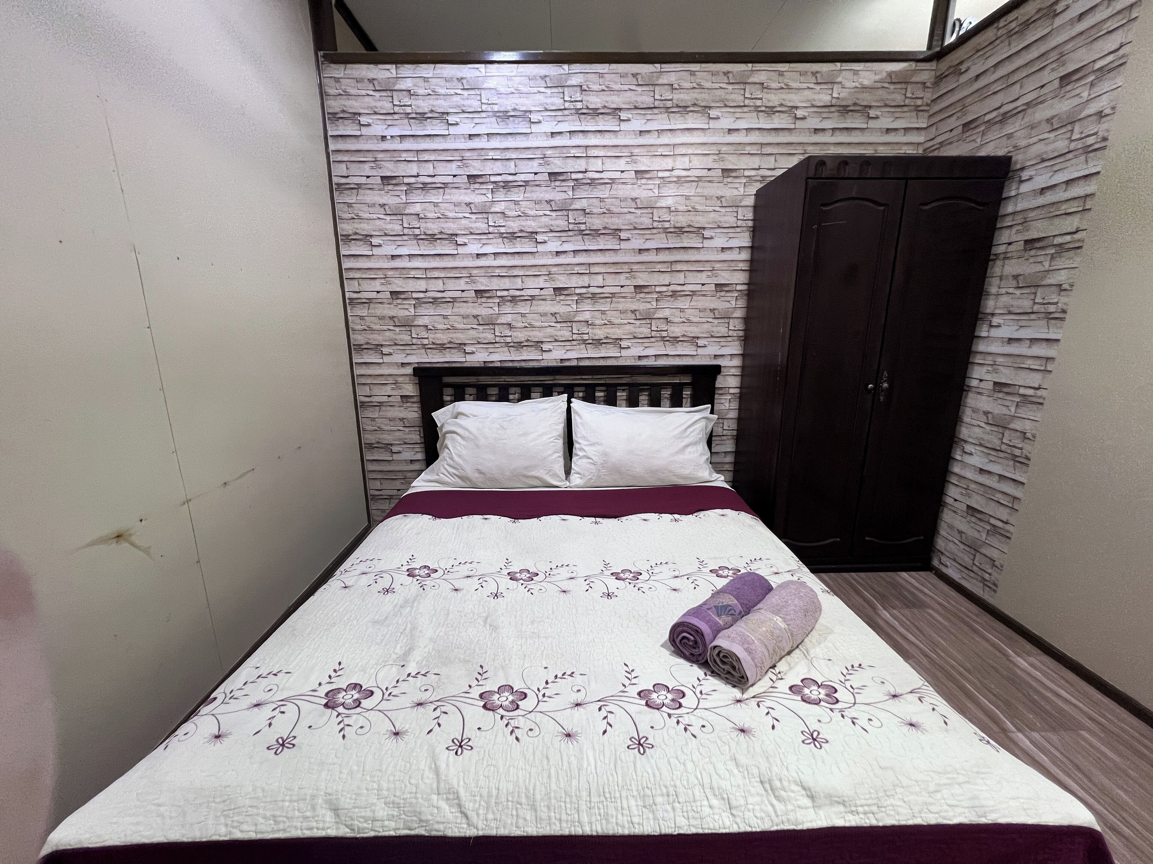 A small, neatly made bed with a floral-patterned bedspread and two pillows, set against a wall with a stone-like texture.