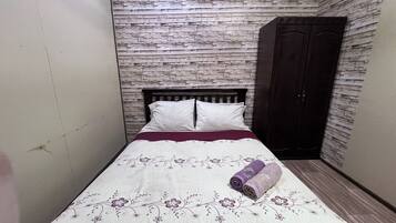 A small, neatly made bed with a floral-patterned bedspread and two pillows, set against a wall with a stone-like texture.