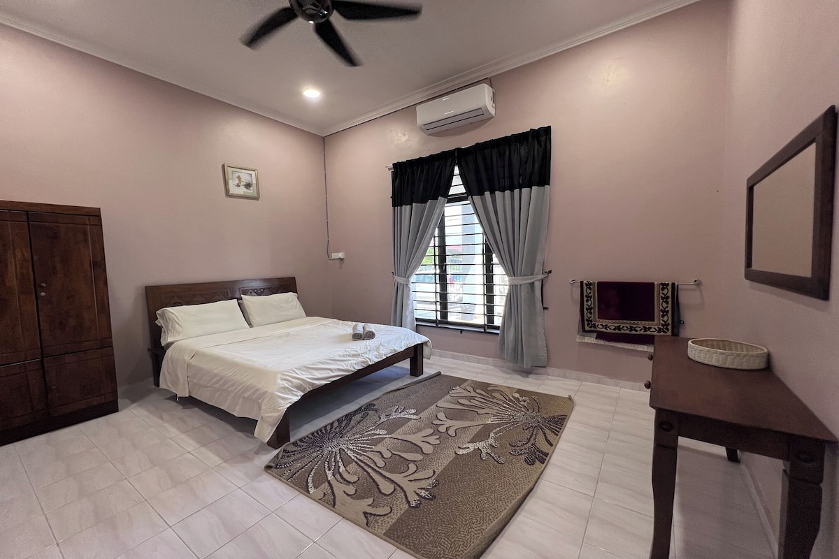A bedroom with a bed, a ceiling fan, an air conditioner, a window with curtains, a bedside table, and a wall-mounted mirror.