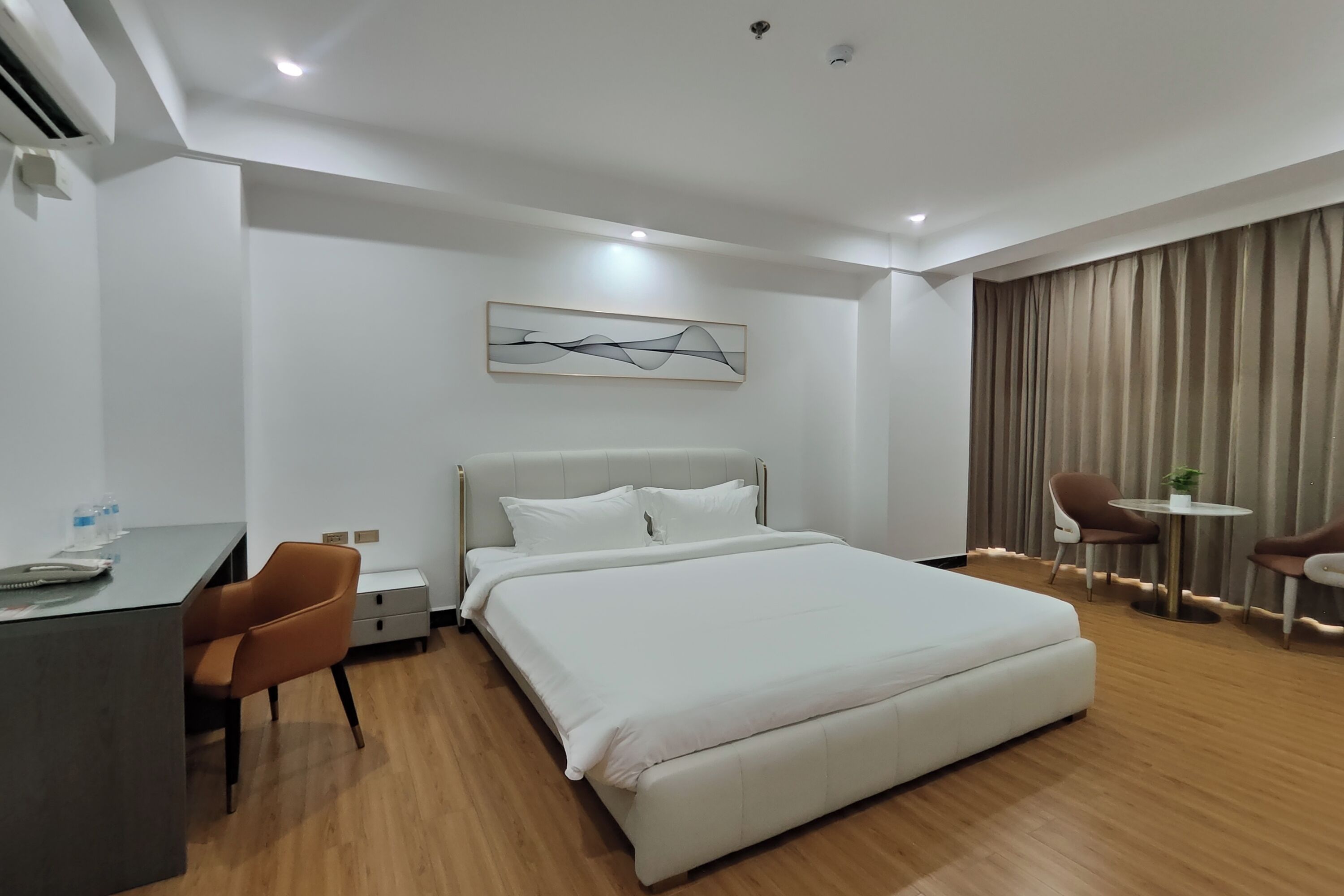 Een hotelkamer met een bed, een bureau en een stoel. Er hangt een schilderij aan de muur en er is een gordijn voor het raam.