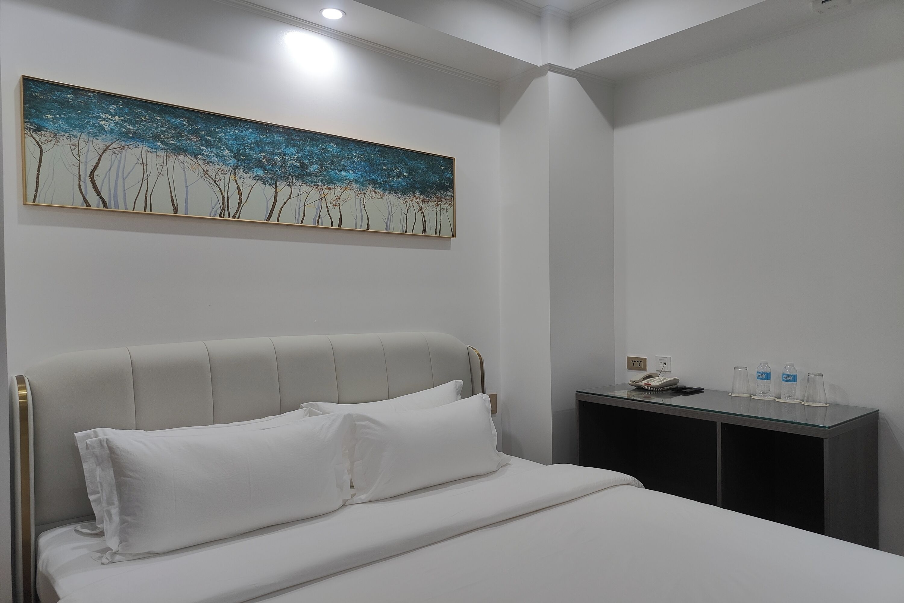 Un letto rifatto con lenzuola bianche, un quadro raffigurante alberi al muro e un tavolino con un telefono e bottiglie d'acqua.