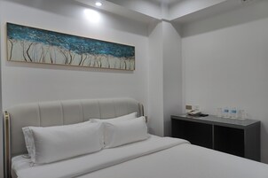 Una cama bien tendida con sábanas blancas, un cuadro con árboles en la pared y una mesita con un teléfono y botellas de agua.