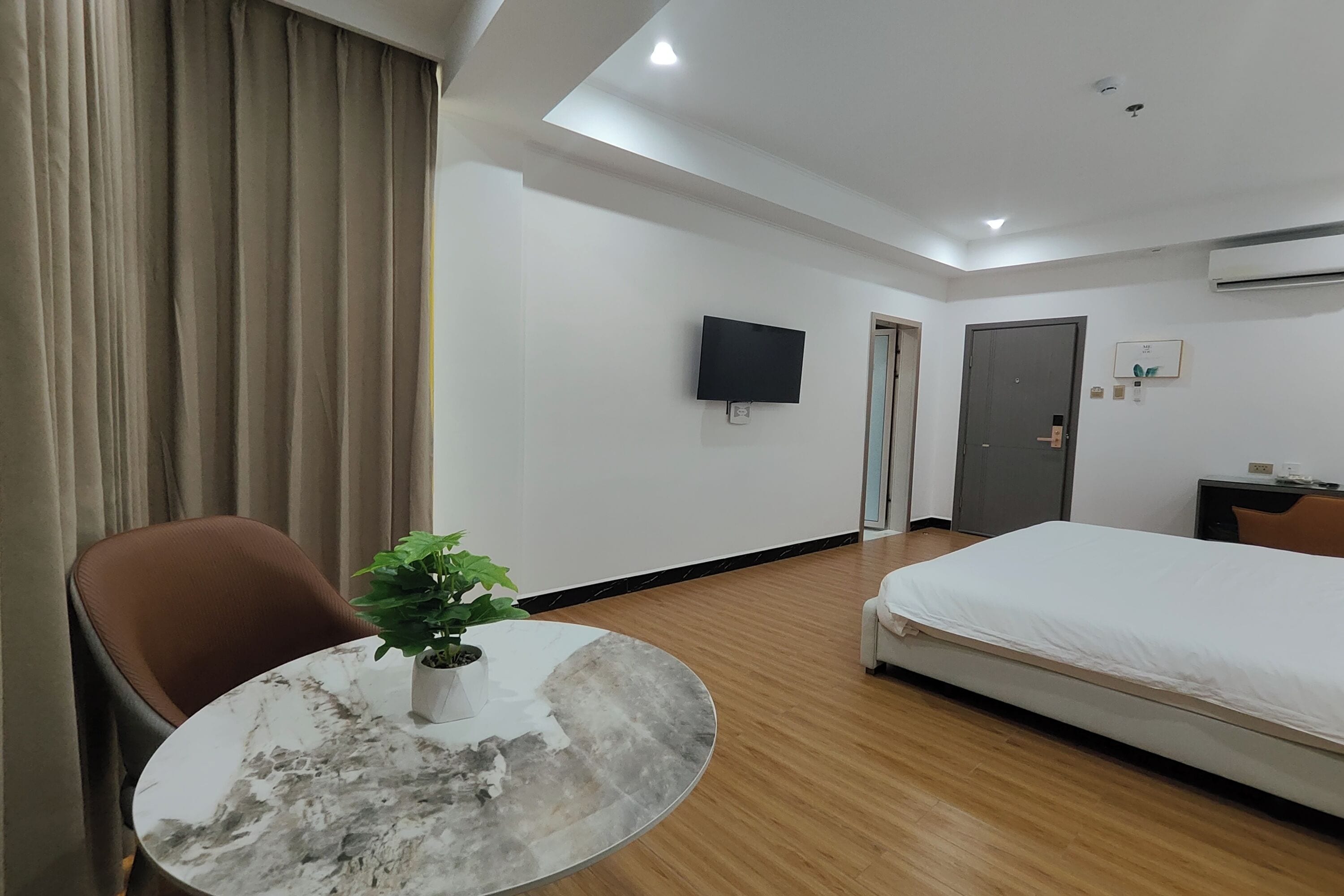 A hotel room with a bed, a TV mounted on the wall, a small table with a plant, and a chair.