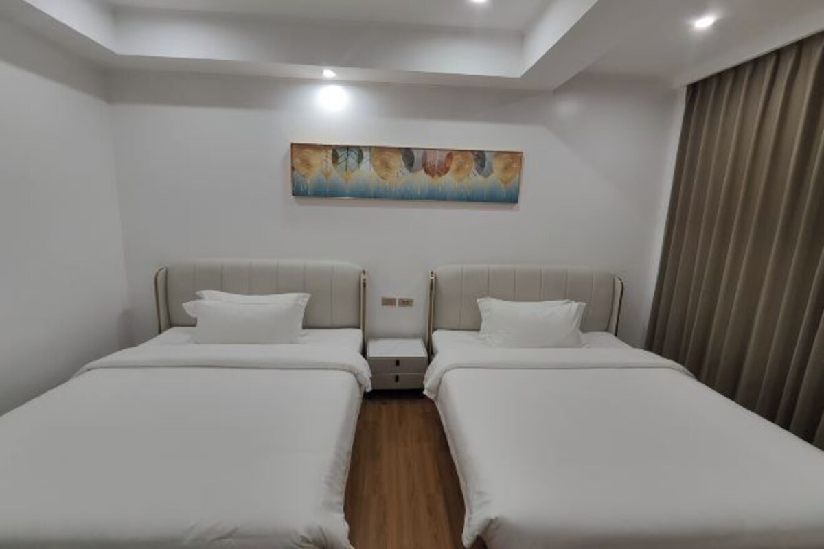 A hotel room with two single beds, a nightstand, a painting on the wall, and a window with curtains.