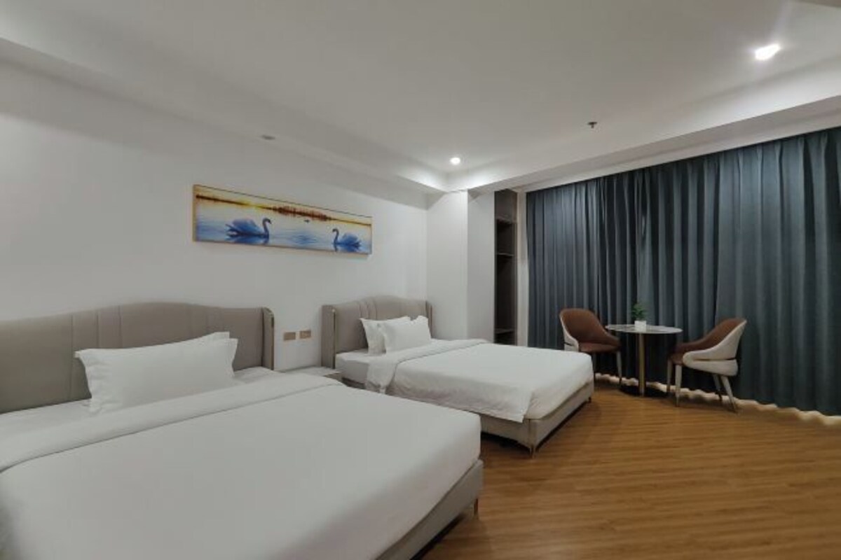 A hotel room with two beds, a painting, a small table with chairs, and a large window with curtains.