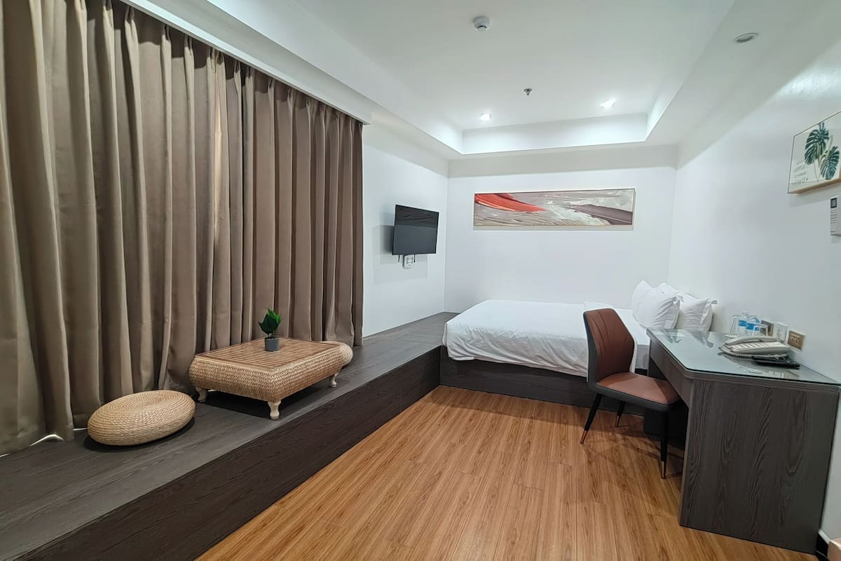 Una habitación de hotel moderna con cama, escritorio, silla, televisor y un amplio ventanal con cortinas.