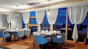Un comedor moderno con mesas dispuestas para dos, sillas azules y grandes ventanas con cortinas blancas.