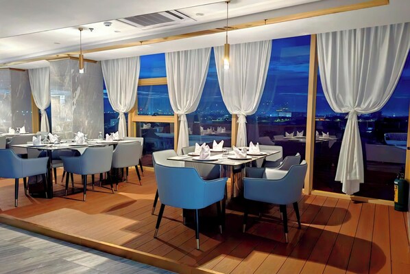 A modern dining area with tables set for two, blue chairs, and large windows with white curtains.