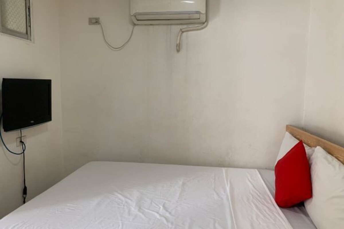 A single bed with white bedding and a red pillow, a wall-mounted air conditioner, and a television on the left side.
