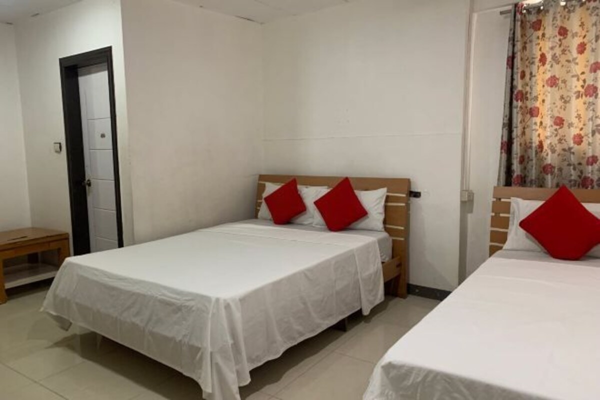 A room with two single beds, each with a white bedspread and red pillows. There is a wooden nightstand beside one bed, and a door with a black frame is visible.