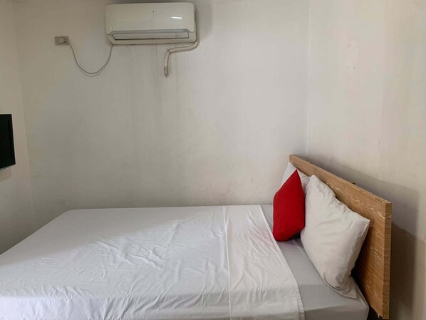 Una cama individual con ropa blanca y una almohada roja, cabecera de madera y un aire acondicionado de pared.