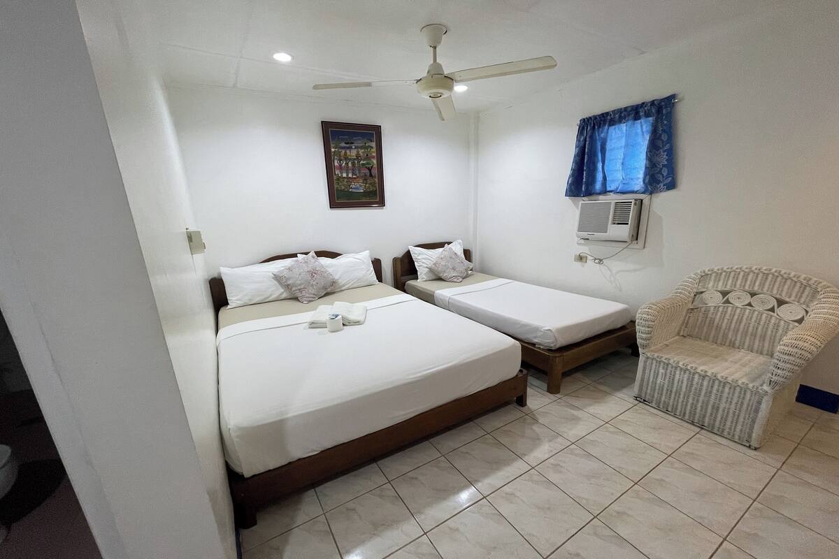 A hotel room with two beds, a ceiling fan, an air conditioning unit, a wicker chair, and a wall-mounted air conditioner.