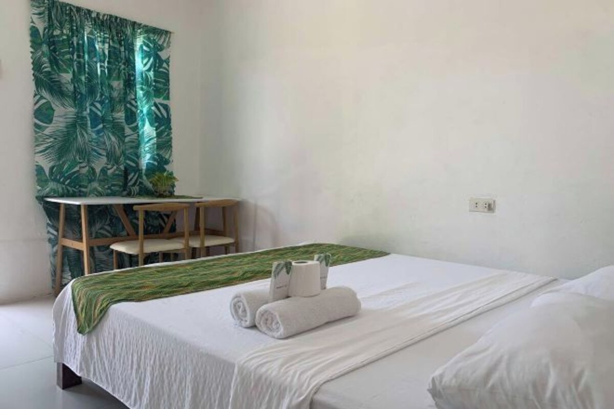 A hotel room with a bed, a desk, and a chair. There is a green curtain with a leaf pattern on the wall.
