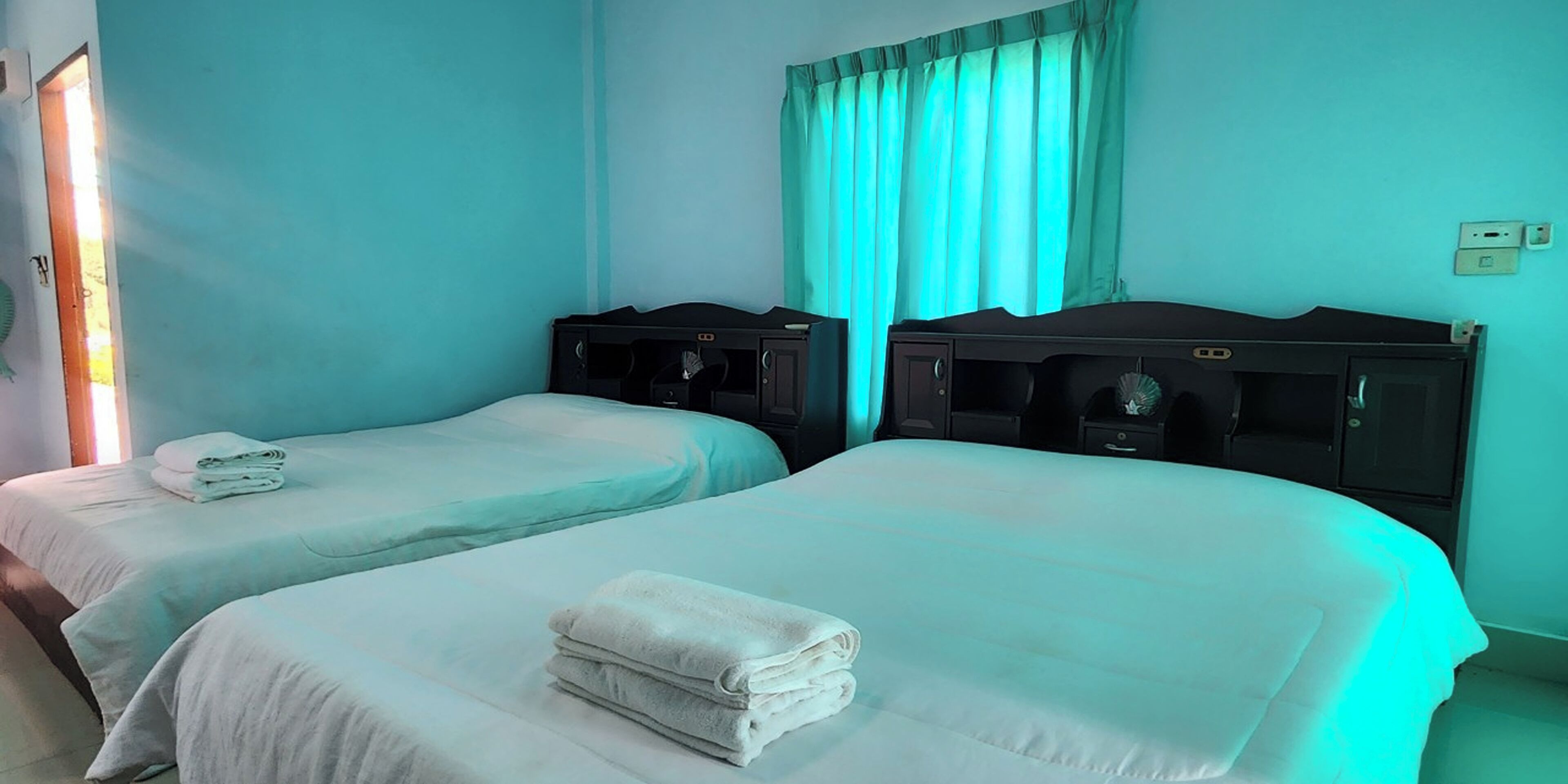 Une chambre d’hôtel avec deux lits, chacun recouvert d’une courtepointe blanche et agrémenté d’une serviette pliée. Les têtes de lit sont en bois foncé et il y a une fenêtre avec des rideaux de couleur turquoise.