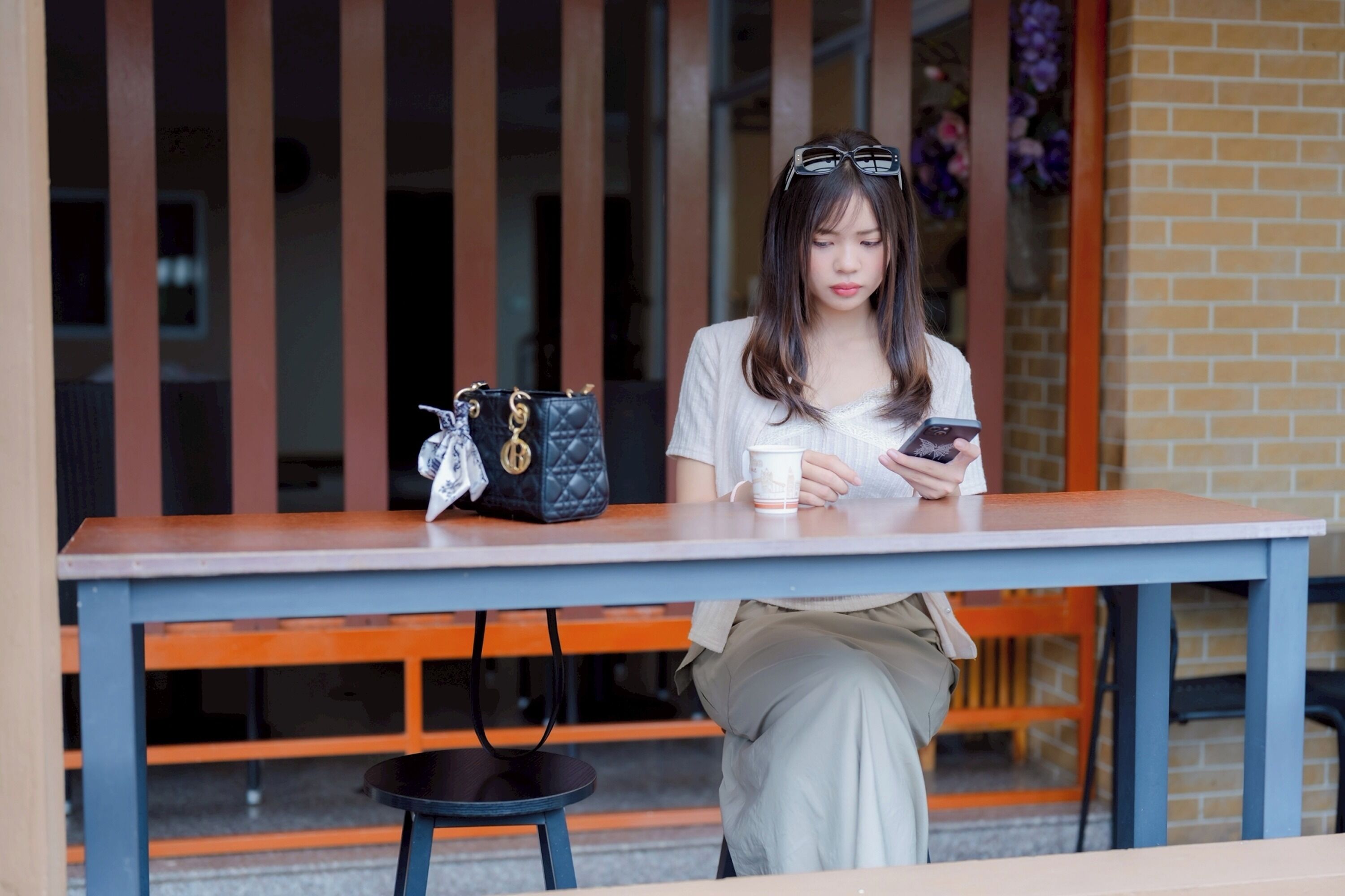 Una persona sentada a una mesa con un teléfono inteligente y una taza, con un bolso de mano y un banco al fondo.