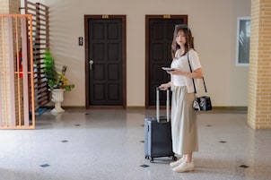 Una persona parada en un pasillo con una maleta, sosteniendo una tableta y vistiendo una blusa blanca y una falda color beige.