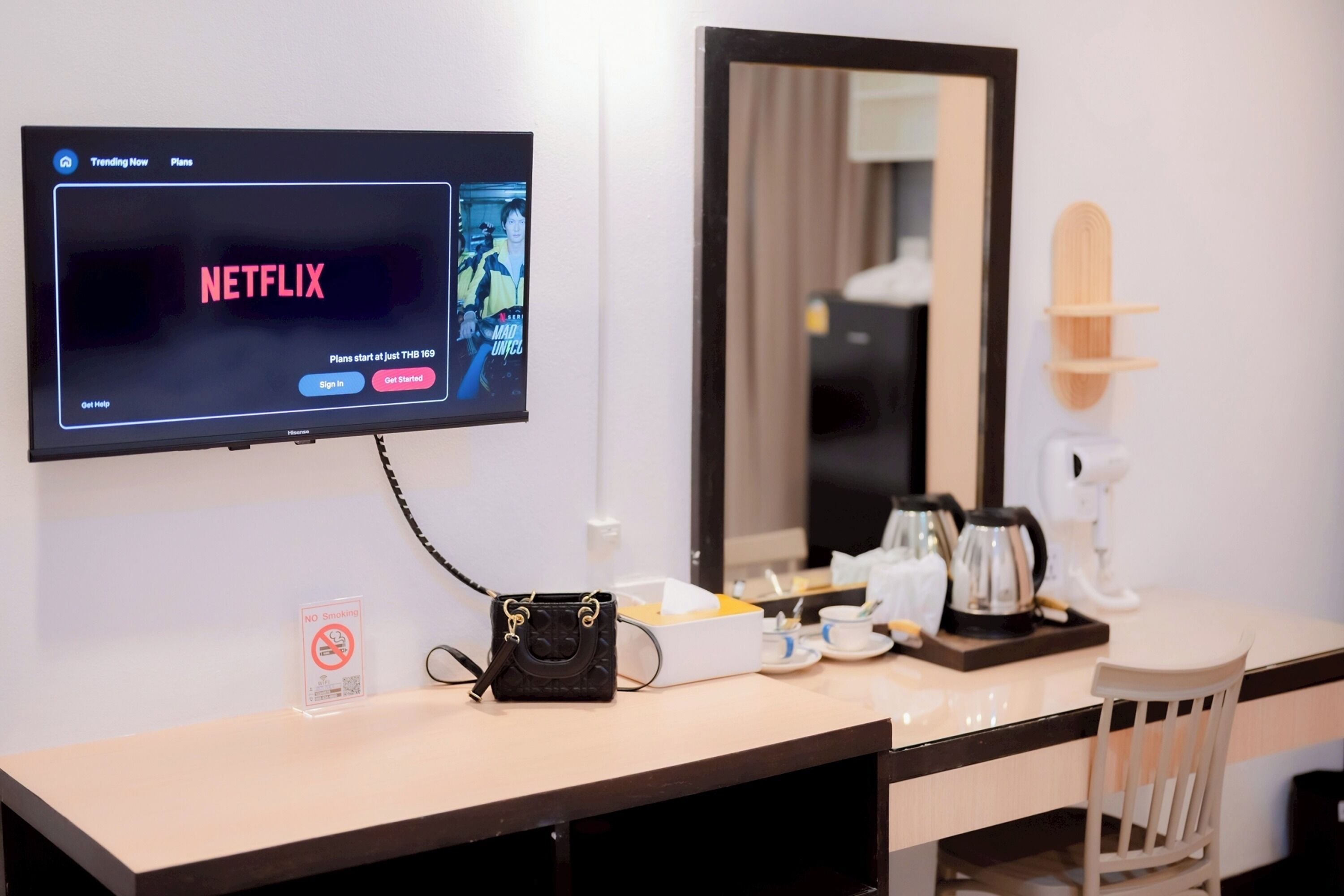 Habitación de hotel con una televisión mostrando la interfaz de Netflix, una cafetera, un hervidor de té, una mesita con un bolso y un cartel de 'NO FUMAR'.