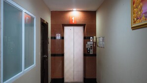 A hallway with a door marked '1' indicating the first floor, a no-smoking sign, an elevator with a control panel, and a wall-mounted phone. - Jt Place Buriram (Buriram)