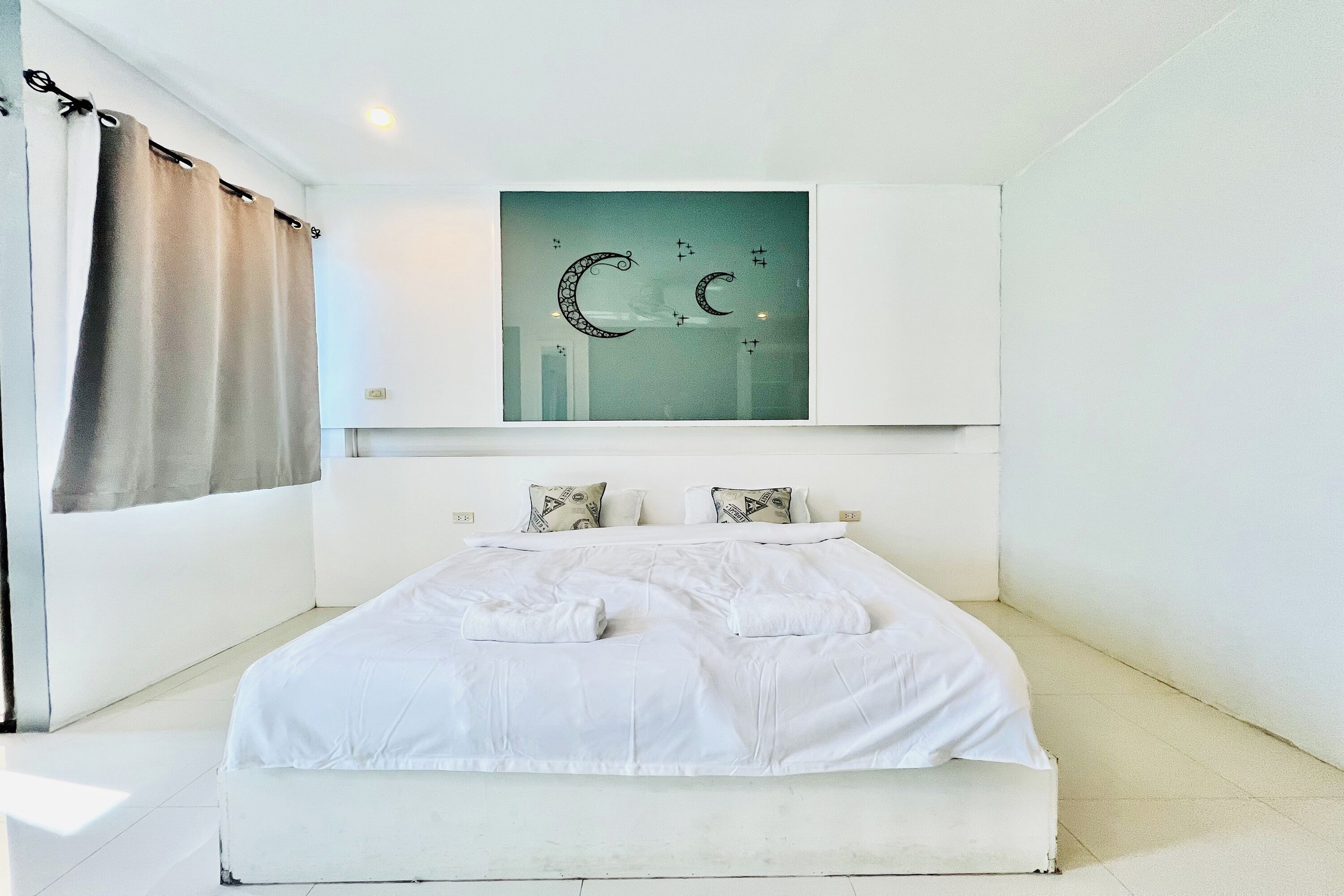 A bedroom with a large bed, white bedding, a wall-mounted artwork of a crescent moon, and a window with curtains.