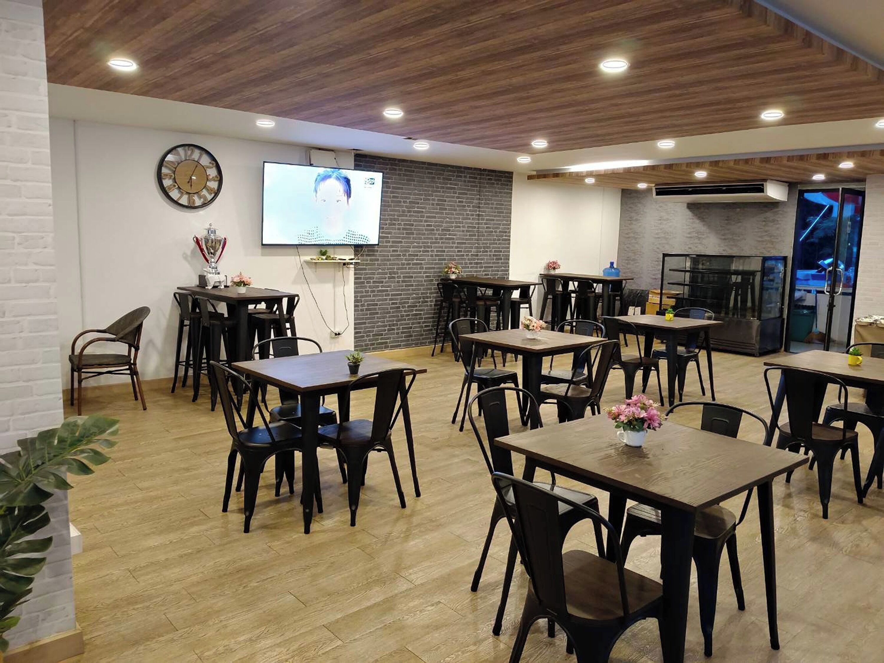 A modern dining area with multiple tables and chairs, a brick wall, a clock, and a television screen.