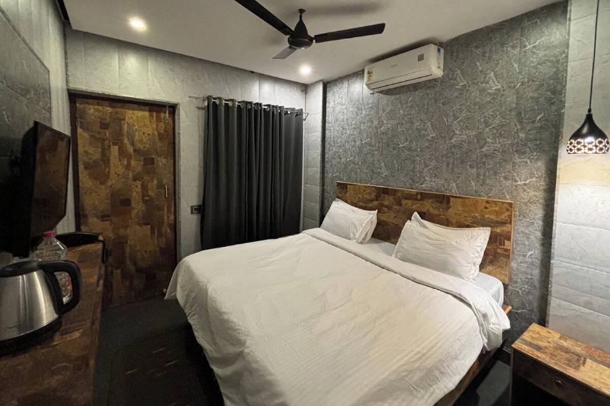 A modern hotel room with a large bed, a ceiling fan, an air conditioning unit, a wooden door, and a small table with a kettle.