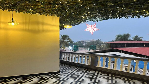 A balcony with a patterned floor, a railing, and a view of palm trees and buildings in the background. - Aariv Comforts Baga (Baga)