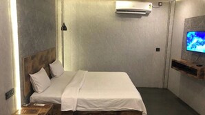 A modern bedroom with a wooden headboard, a white bedspread, an air conditioning unit, a bedside table with a lamp, and a mounted TV. - Aariv Comforts Baga (Baga)
