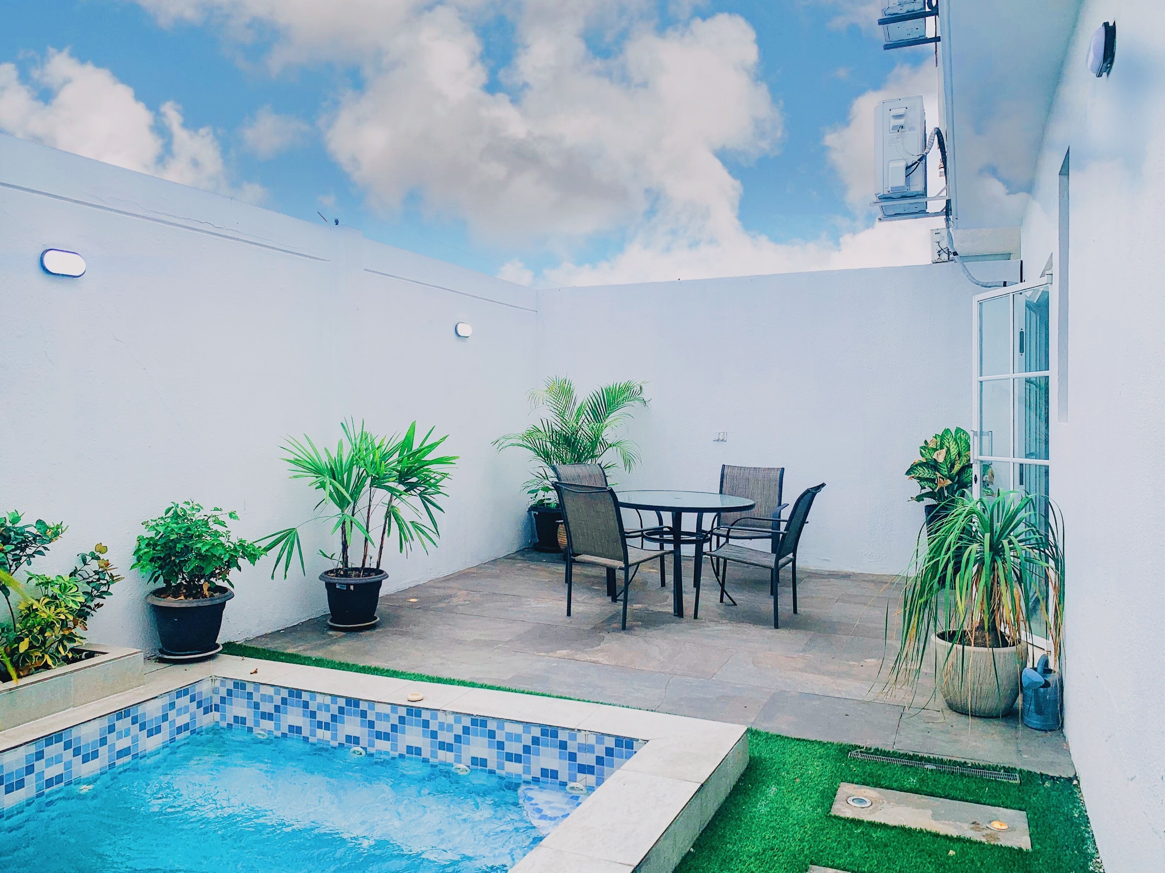 A small outdoor area with a pool, a table with chairs, and several potted plants.