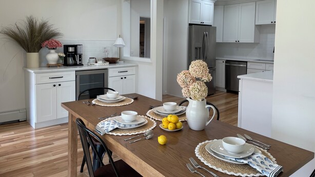 Una cocina moderna con armarios blancos, un refrigerador de acero inoxidable y un comedor con una mesa de madera dispuesta para cuatro.