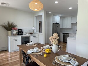 A modern kitchen with white cabinets, a stainless steel refrigerator, and a dining area with a wooden table set for four. - NEW! Updated rare 9 bedroom, sleeps 18-20 & short walk to Roger Wheeler Beach! (Narragansett)