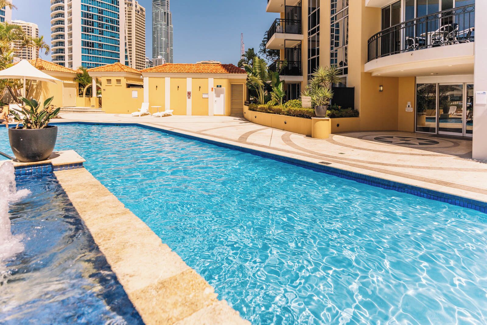 A swimming pool with a water fountain, surrounded by a building with balconies and a poolside area with lounge chairs.