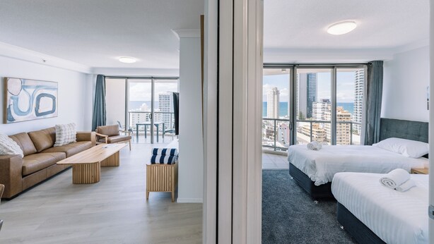 A modern hotel room with a large window, a sofa, a coffee table, and two beds. - Resort Style Chevron Apartments (Surfers Paradise)